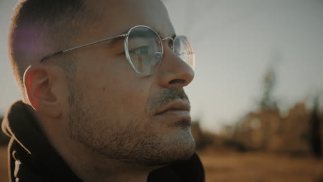 Inspired young Caucasian man wearing glasses breathing fresh air out in nature at sunset or sunrise