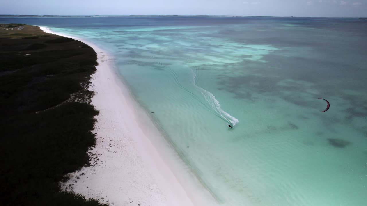A kitesurfer rides across clear turquoise waters near a white sandy beach in Los Roques, Venezuela