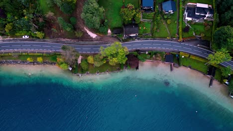 An-aerial-view-of-a-winding-road-lined-with-trees-and-houses-leading-to-a-blue-and-green-lake,-with-a-clear-blue-sky-above