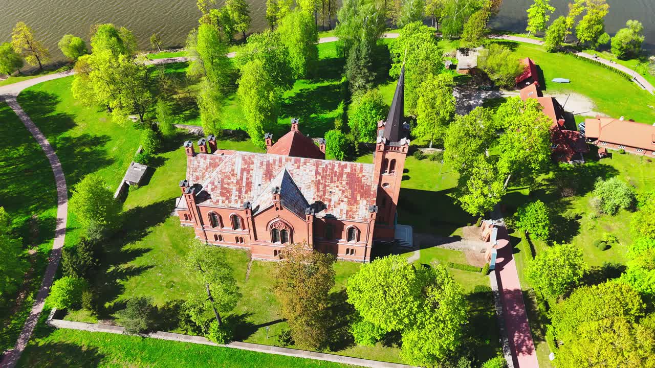 A red brick church with a sharp steeple rests near a lakeshore, surrounded by walking paths, trees, and vibrant green parkland under clear spring skies.