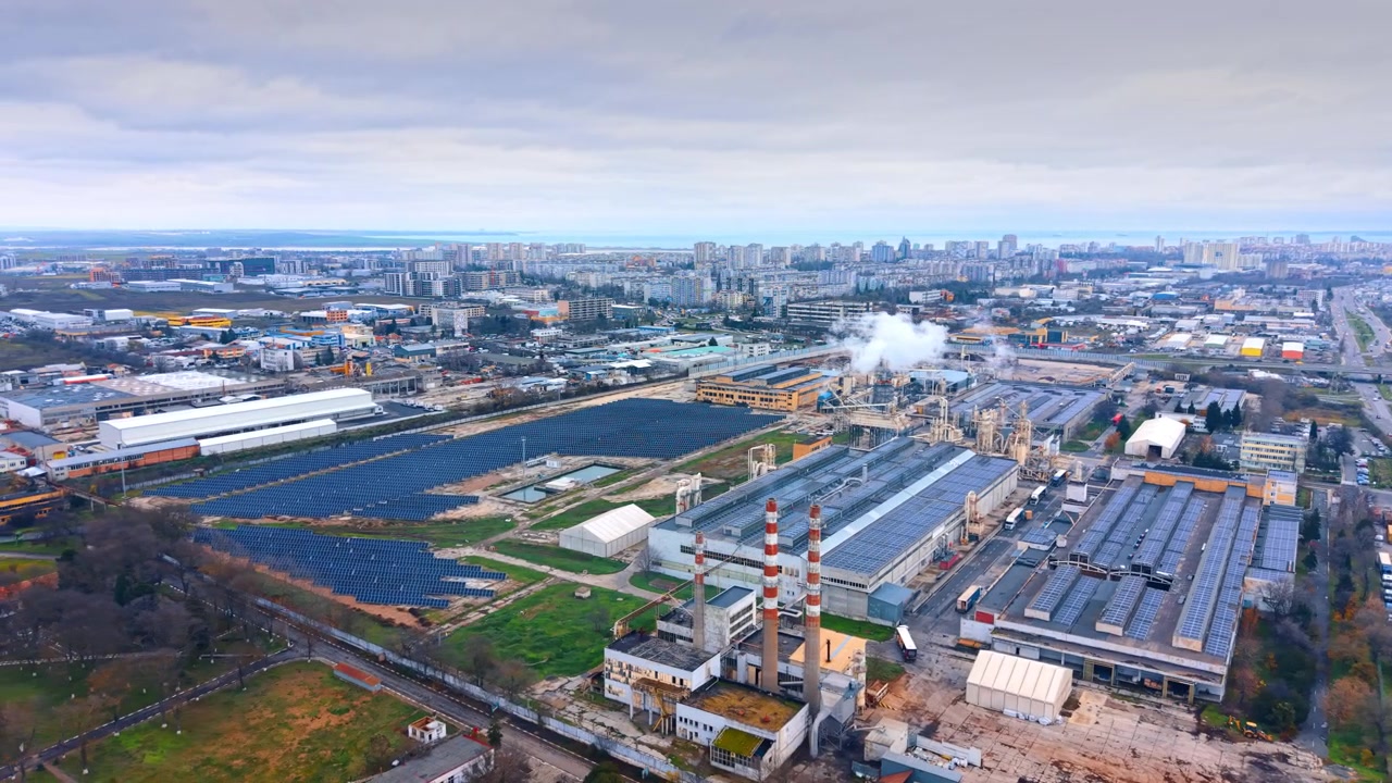 Güneş panelleri ve şehir manzarası olan sanayi fabrikasının havadan görünümü. Güneş paneli dizisi, fabrika çatıları ve deniz kenarında uzakta bir şehir içeren büyük bir sanayi tesisinin drone görüntüsü