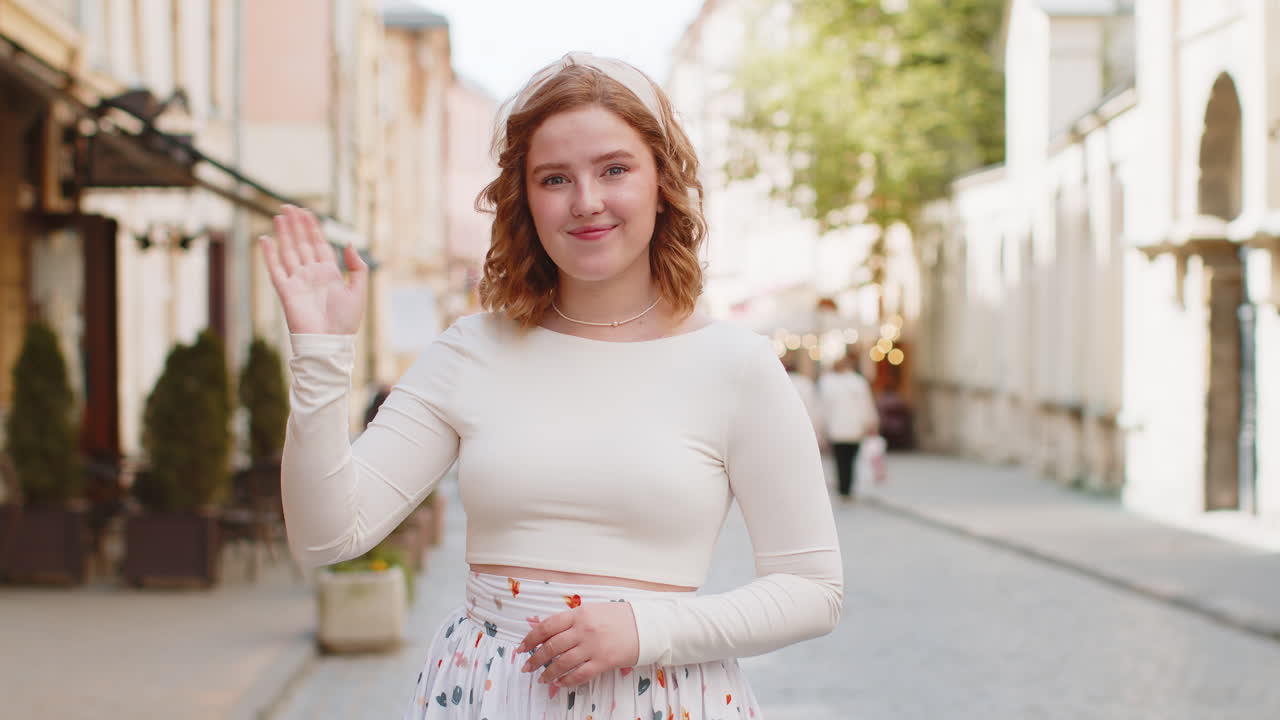 Redhead woman smiling friendly at camera waving hands hello hi greeting or goodbye in city street