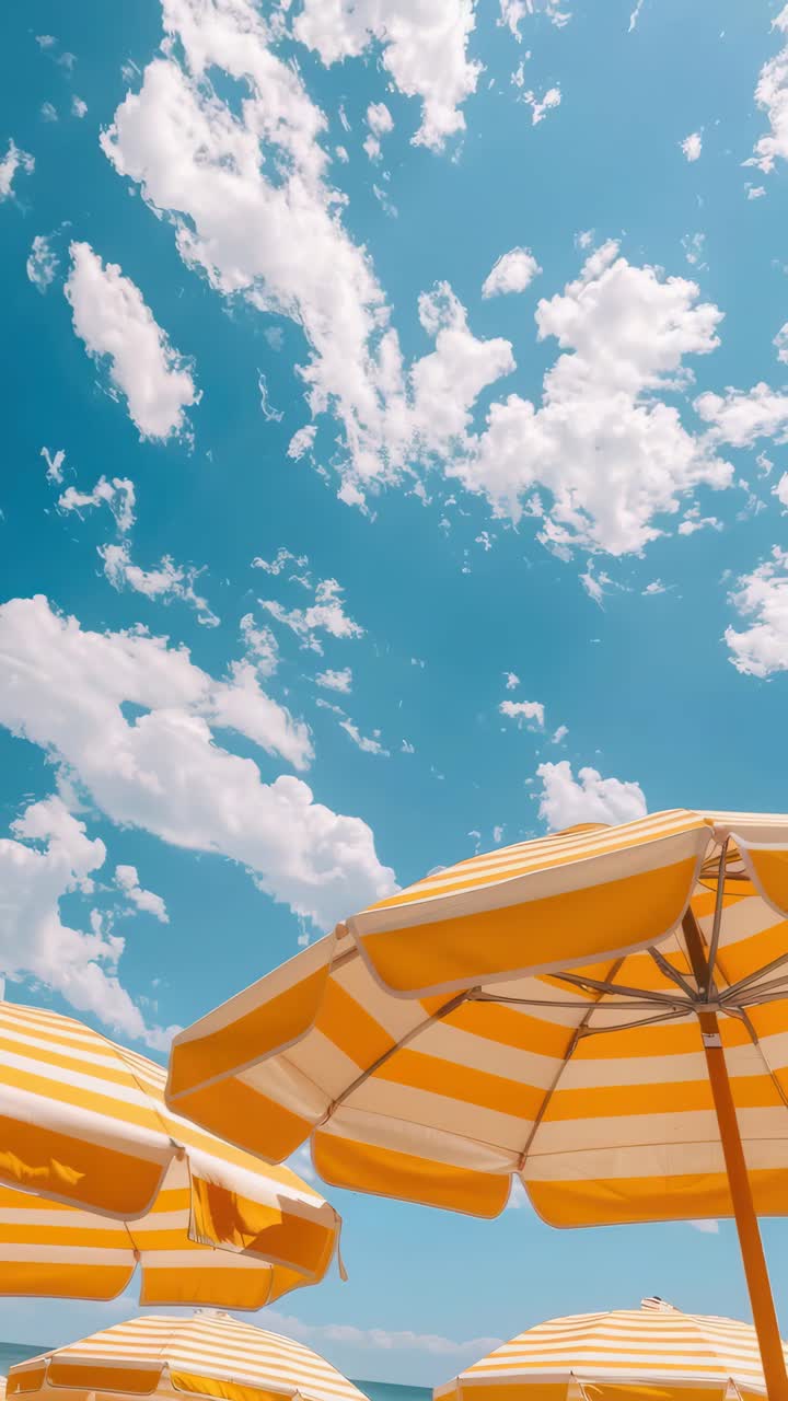 Low-angle video frame capturing yellow-striped beach umbrellas against a bright blue sky with fluffy