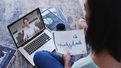african american woman holding a document having a video call with male colleague on laptop