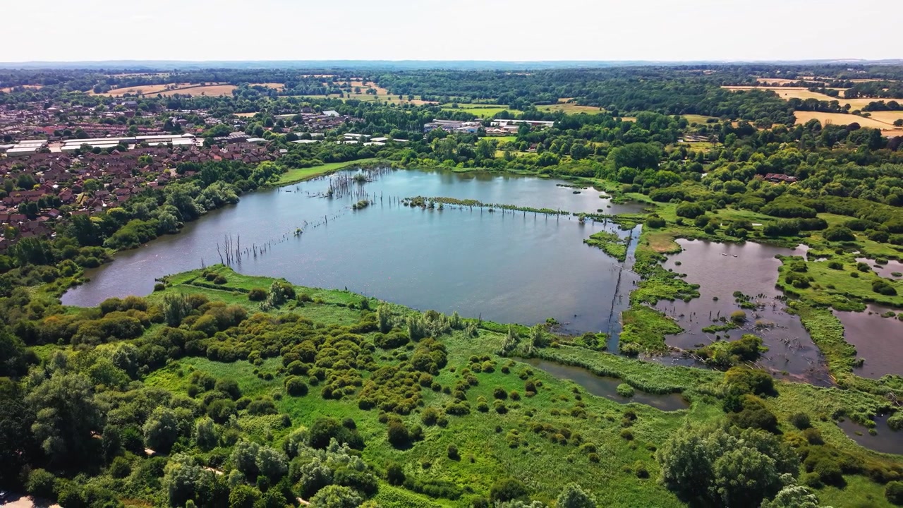 Drone trekker seg tilbake fra Fishlake Meadows' åpne innsjø, og avslører våtmarkskratt og Romseys hustak i det fjerne