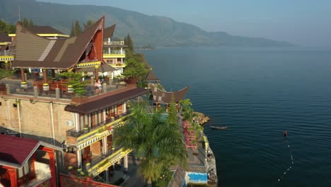 Flyover-drone-footage-of-lakeside-resorts-with-traditional-Batak-roofs-on-Lake-Toba-in-North-Sumatra,-Indonesia