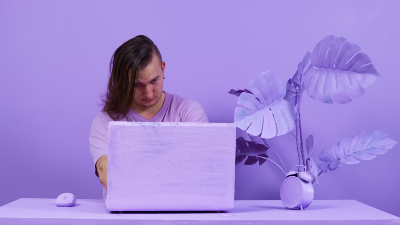 Young man with long hair finishes work closing laptop at table near pot-plant. Office with interior painted in purple color. Life in fantasy world