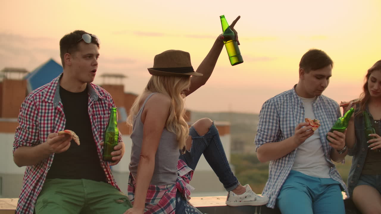 Young russian people sit on the roof and drink beer with pizza. They enjoy a beautiful sunset and time with their mate. They raise their hands up and clink glasses of beer.