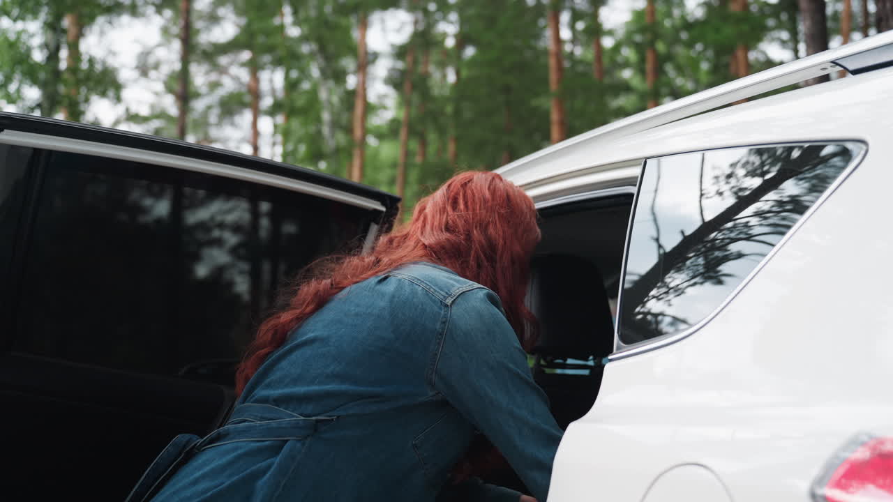 빨간 머리 여성이 데님 재킷을 입고 숲에 주차된 흰색 차에서 담요를 꺼내 피크닉을 준비하는 모습의 낮은 뷰예요. 초록색 나무와 따뜻한 햇살로 가득한 평온한 야외 순간이에요.