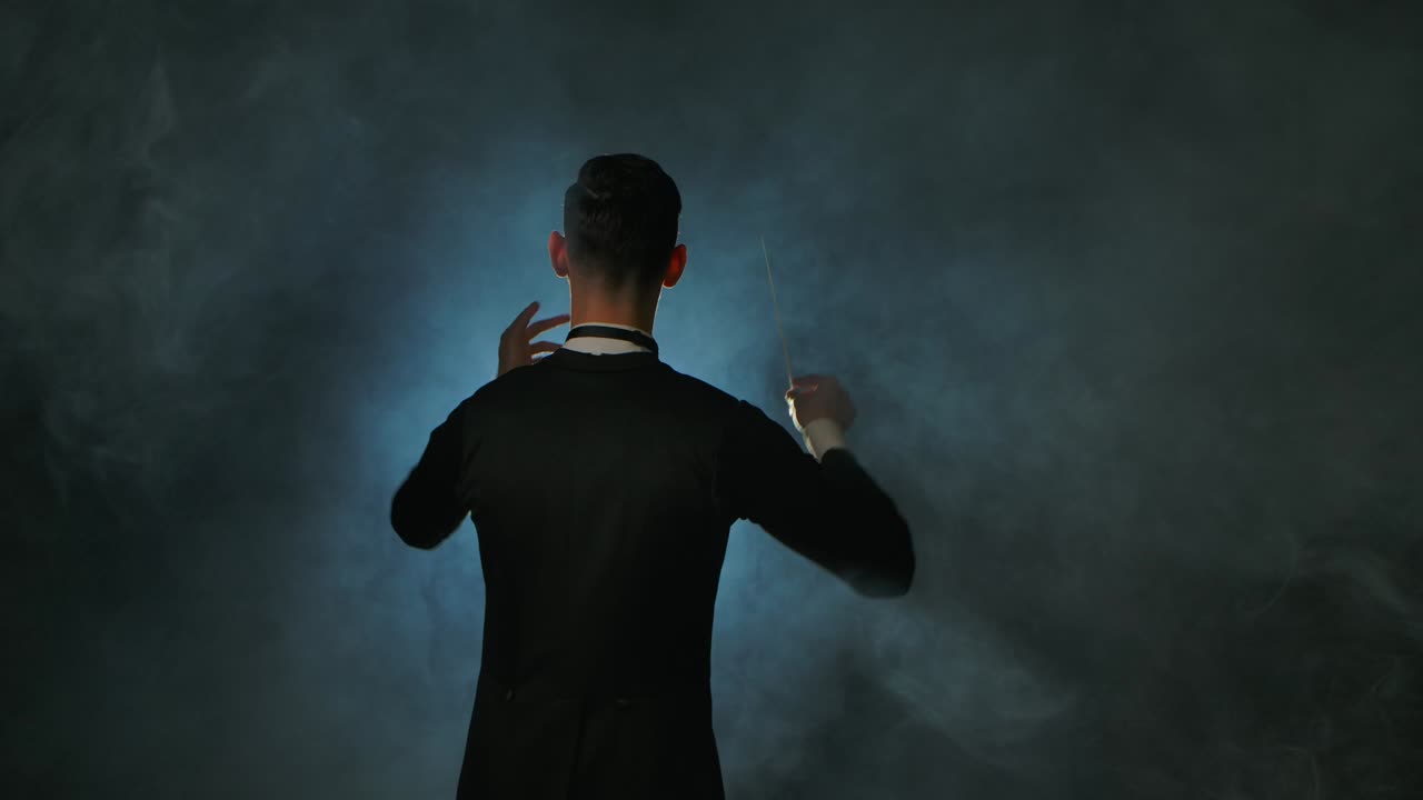 심포니 오케스트라 지휘자: 마에스트로는 검은 연기 배경과 백라이트에서 막대기의 움직임으로 음악가들을 지휘하고 있으며, 가시적인 몸짓으로 음악 공연을 지휘하고 지휘하고 있습니다.