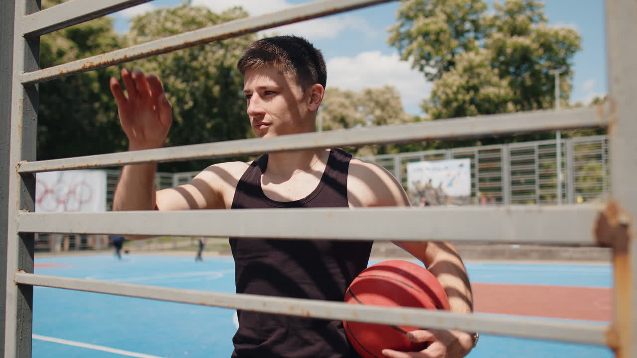 Portrait of athletic serious man playing basketball posing alone stands behind grid on urban court