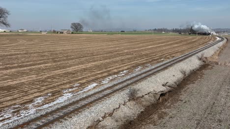 A quiet rural area showcases train tracks winding through freshly plowed fields. Smoke gently rises in the distance, of an approaching steam train
