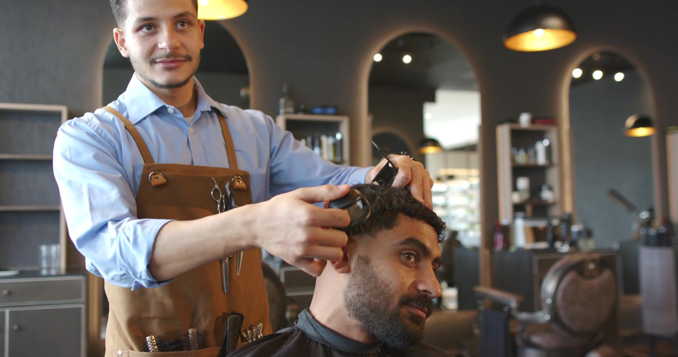 Barbeiro e cliente, de avental, cortando com máquina enquanto o cliente inclina a cabeça, criando um degradê na barbearia