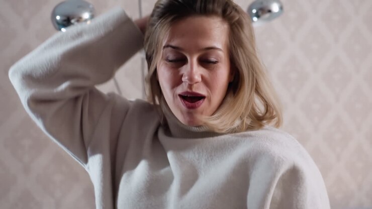 Donna con dolcevita bianca che scuote la testa a occhi chiusi in interni, mostrando movimento espressivo, energia giocosa, umore spensierato, stile di vita casual e atmosfera accogliente in un elegante interno domestico