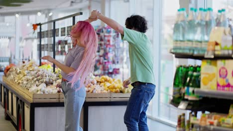 una chica feliz con el cabello rosa en vaqueros y un chico moreno en una camiseta verde están bailando cerca del mostrador en un supermercado durante sus compras