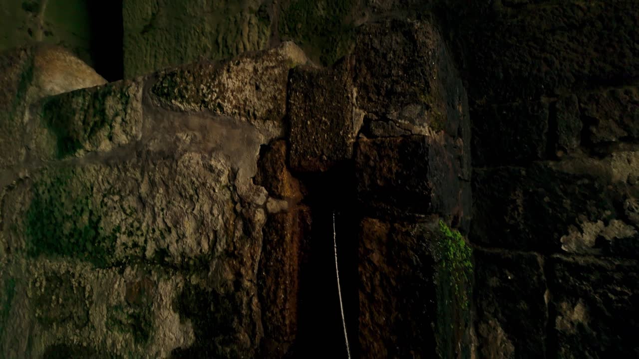 Muri in pietra scuri e robusti con superfici profondamente texturizzate, caratterizzati da una piccola apertura ad arco. L'interno scarsamente illuminato suggerisce un antico passaggio sotterraneo o le profondità di una fortificazione storica.