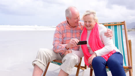 vista frontal de una pareja de ancianos caucásicos usando un teléfono móvil y sentados en una tumbona en una playa 4k