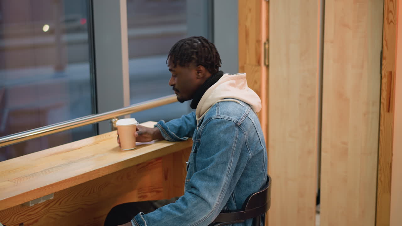Young man in denim jacket and beige hoodie sits comfortably in modern lounge sipping hot coffee from paper cup, facing window with city lights and blurred motion outside