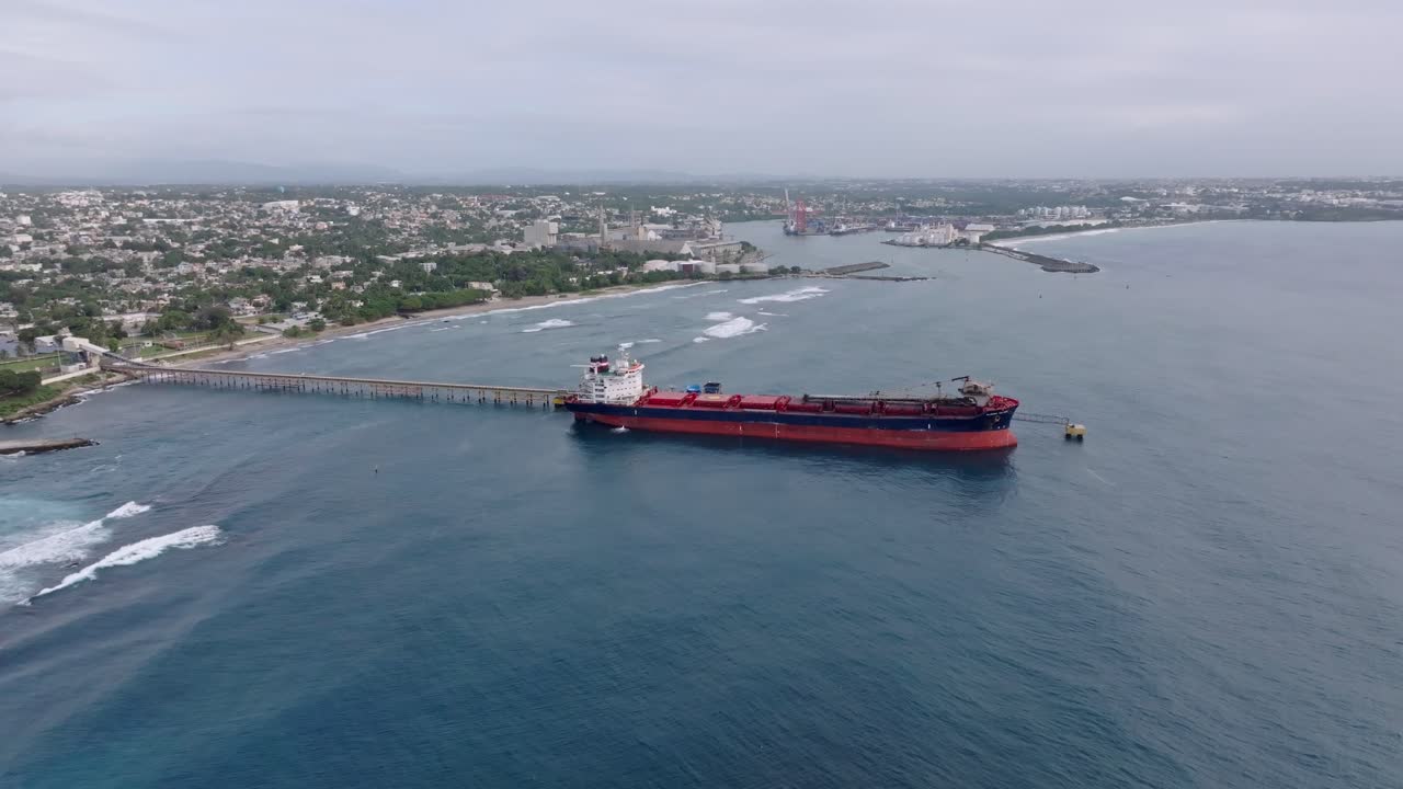 Docking Oil Tanker delivery on port of haina with pipeline. Aerial backwards wide shot. Caribbean sea during cloudy day.