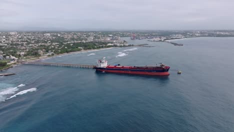 docking oil tanker delivery on port of haina with pipeline. aerial backwards wide shot. caribbean sea during cloudy day.