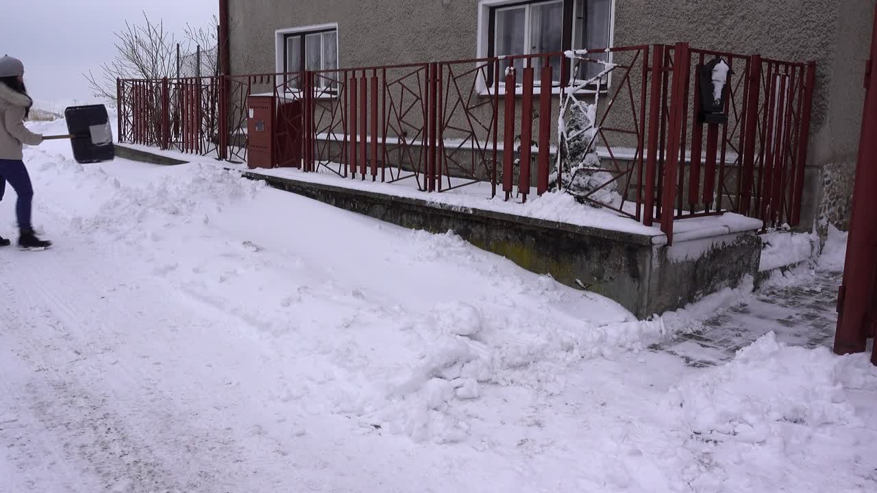 집 밖에서 눈을 삽질하는 겨울 옷을 입은 젊은 유행 여성