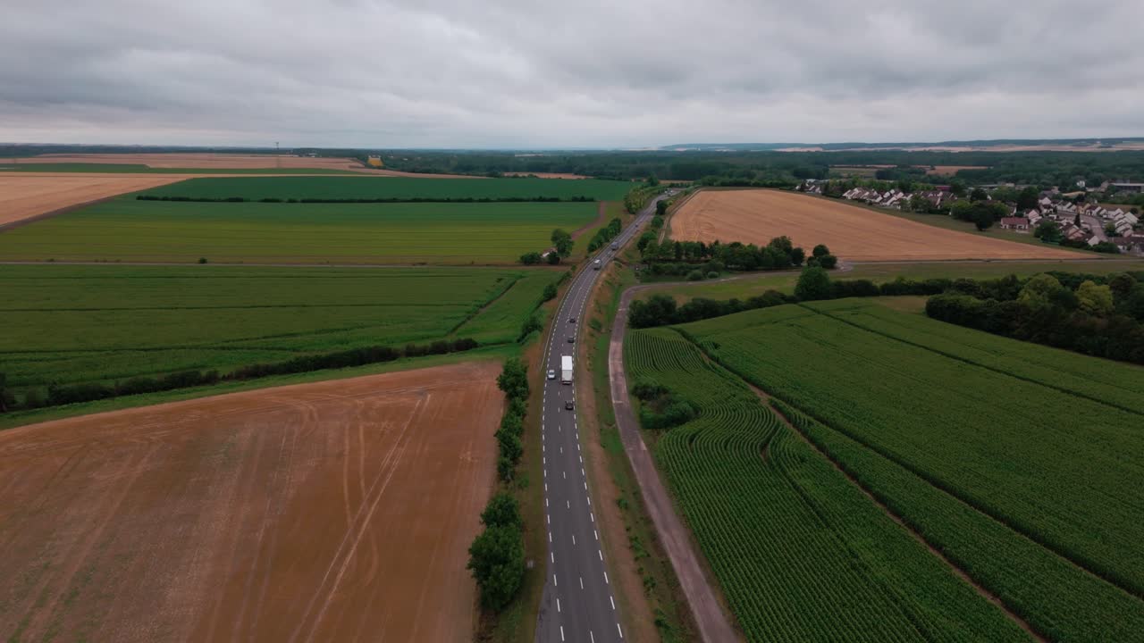 High angle, static aerial drone view of a road with cars and trucks driving through vast green and brown agricultural fields under a cloudy sky in rural France