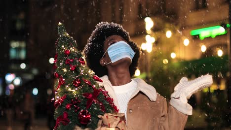 vista de cerca de una alegre mujer afroamericana con máscara facial sosteniendo un árbol de navidad y jugando con la nieve en la calle en navidad