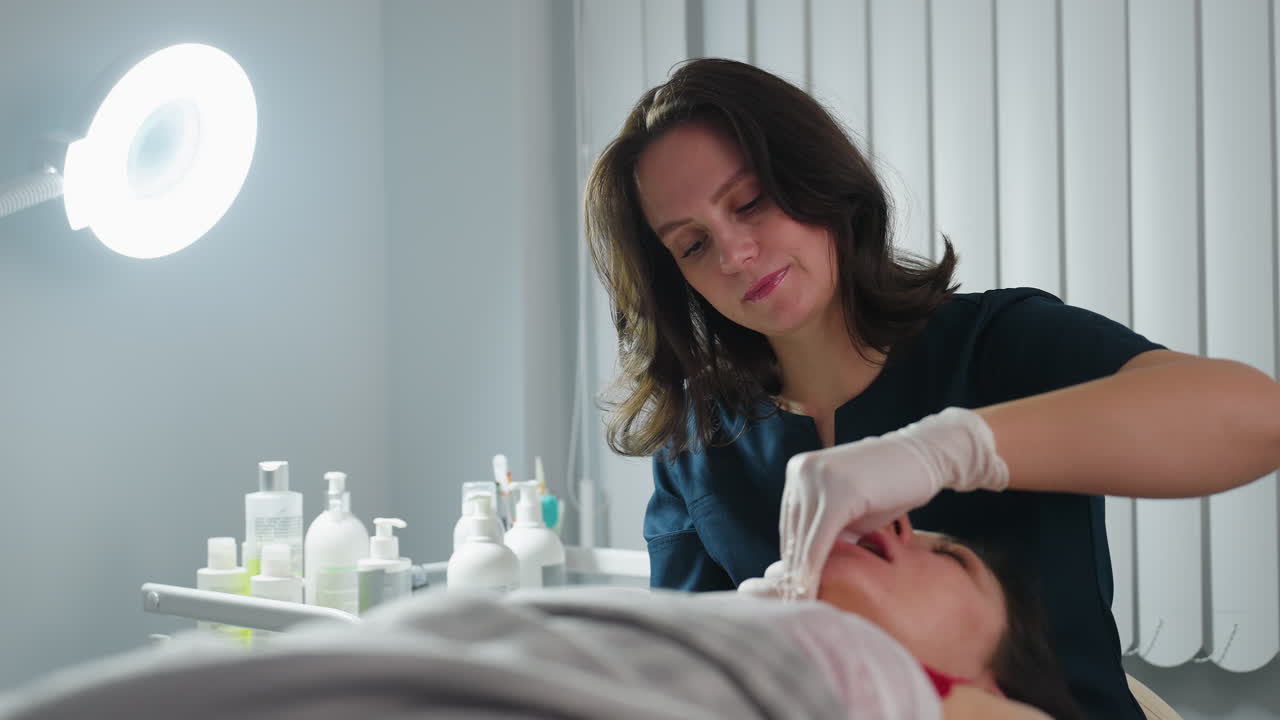 Close up of aesthetician wearing disposable gloves dipping fingernail inside mouth of client while other hand cradles jawline smoothly during spa session in clinic with bottles and equipment nearby