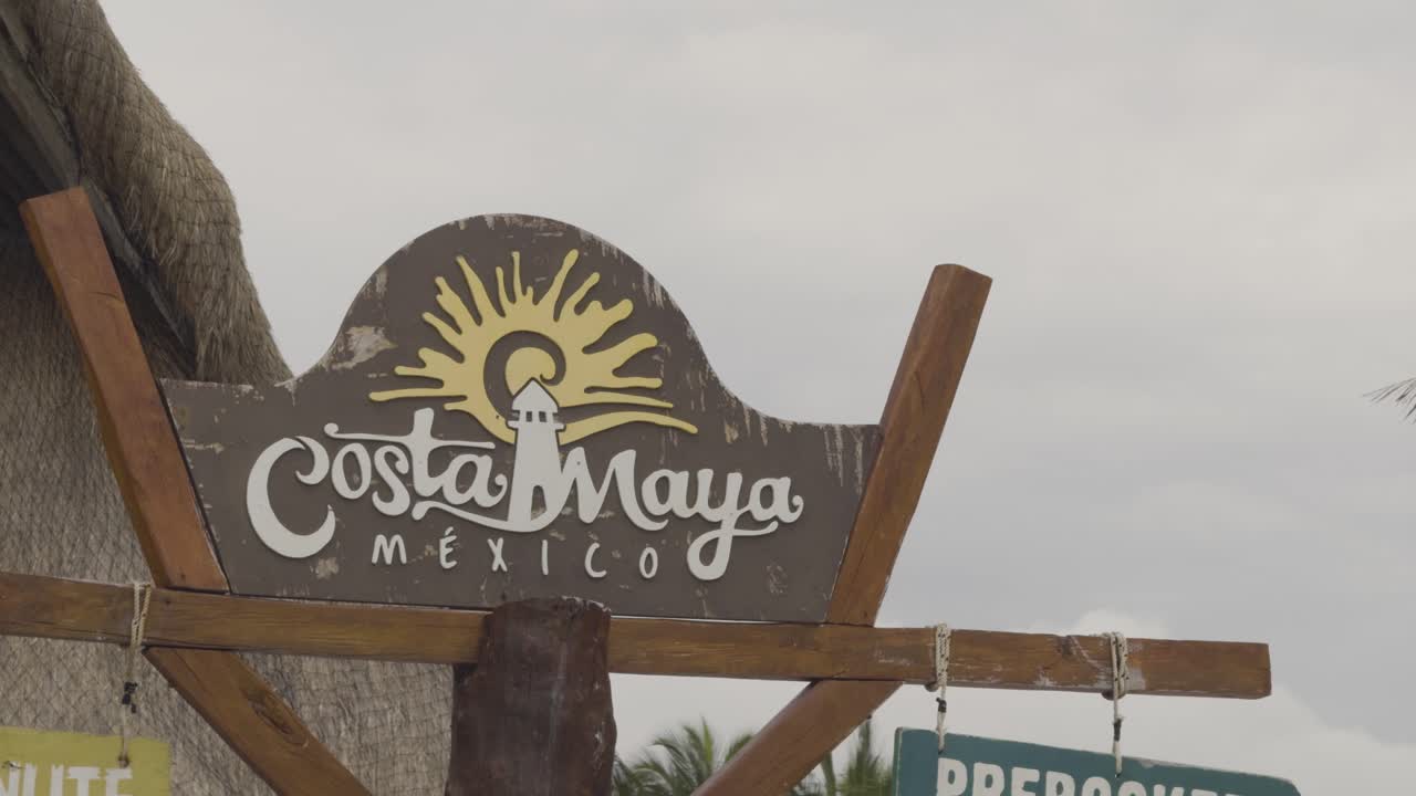 Closeup of Costa Maya sign beneath an overcast sky, with palm trees swaying in the breeze.