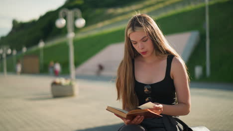 mujer sentada al aire libre en un banco con las piernas cruzadas, volviendo una página de su libro y observando pensativamente un pedazo de papel, luego colocándolo de nuevo, el fondo presenta personas borrosas caminando