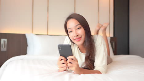 face close-up on asian woman watching video on mobile phone holding smartphone with both hands while comfortably lying on a bed in a hotel room