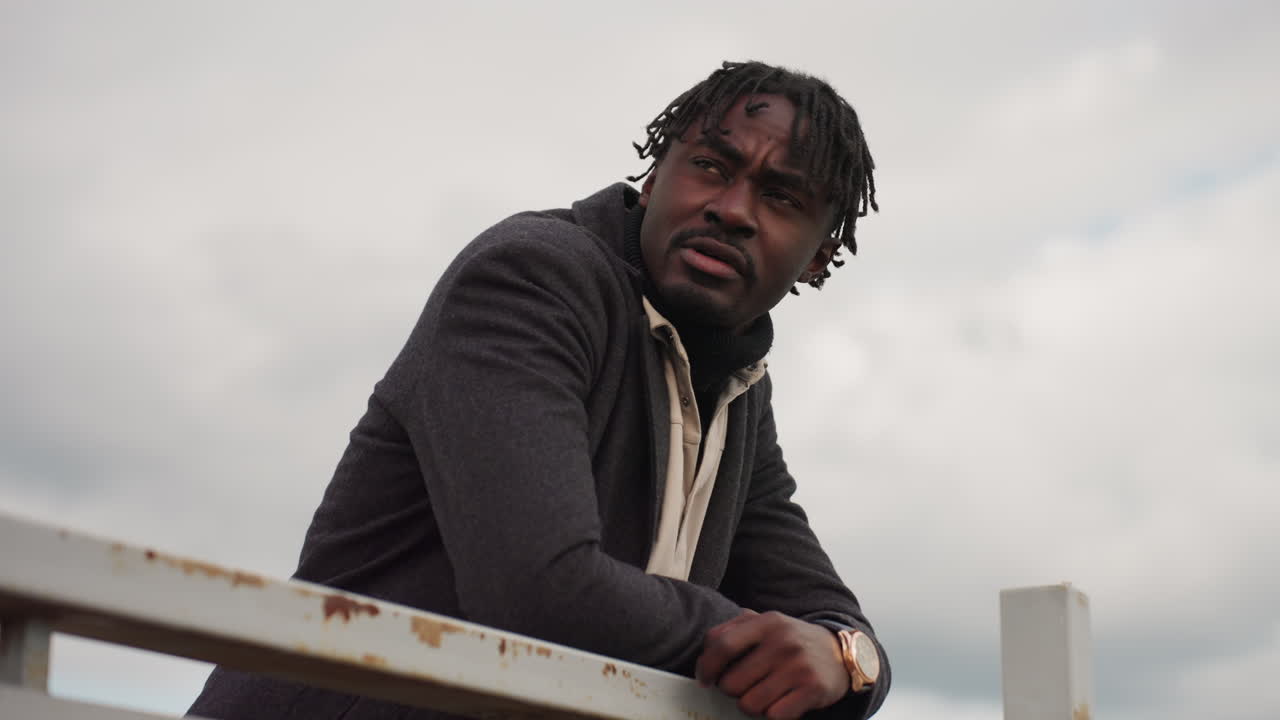 Handsome young man leans on iron handle and looks around outdoors, wearing coat under cloudy sky, showing calm thoughtful mood and modern style while embracing quiet atmosphere of natural environment