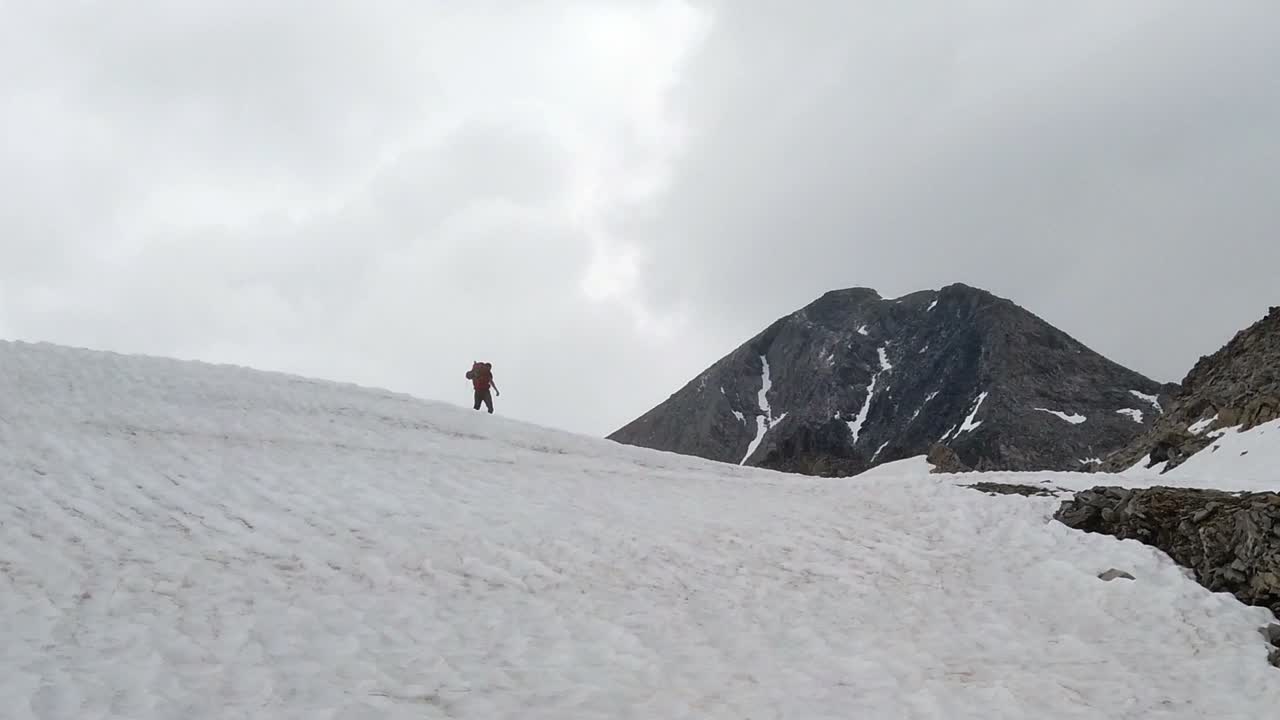 Hiker with backpack slowly walking over snow field. He is careful not to fall. High mountains and cloudy sky in background