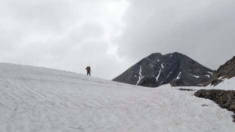 hiker with backpack slowly walking over snow field. he is careful not to fall. high mountains and cloudy sky in background
