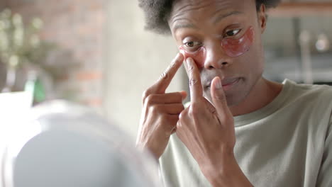 african american man sitting on sofa, applying under-eye patches at home, slow motion