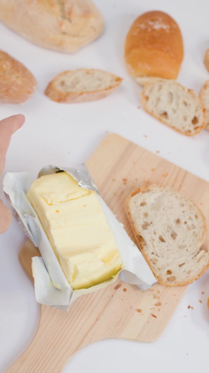 Inquadratura dall'alto di vari tipi di pane e marmellata su sfondo bianco, mentre il burro viene scartato