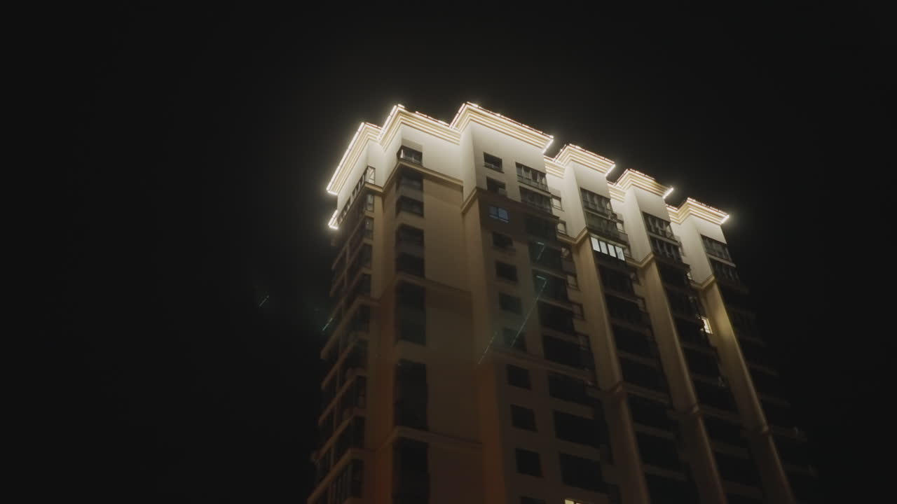 corona de torre nocturna vista desde un coche, perspectiva en primer plano que se extiende hacia arriba a lo largo de la fachada iluminada, luces ornamentadas en la azotea que brillan, ventanas que reflejan el resplandor de la calle, movimiento vertical dramático, cinematográfico