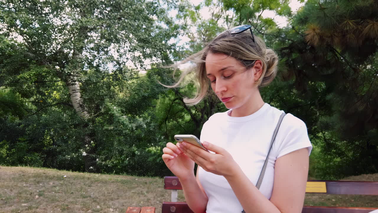 A young woman in a white top sits on a bench in a park, focused on her phone. Surrounding trees provide shade as she enjoys the outdoors on a bright day