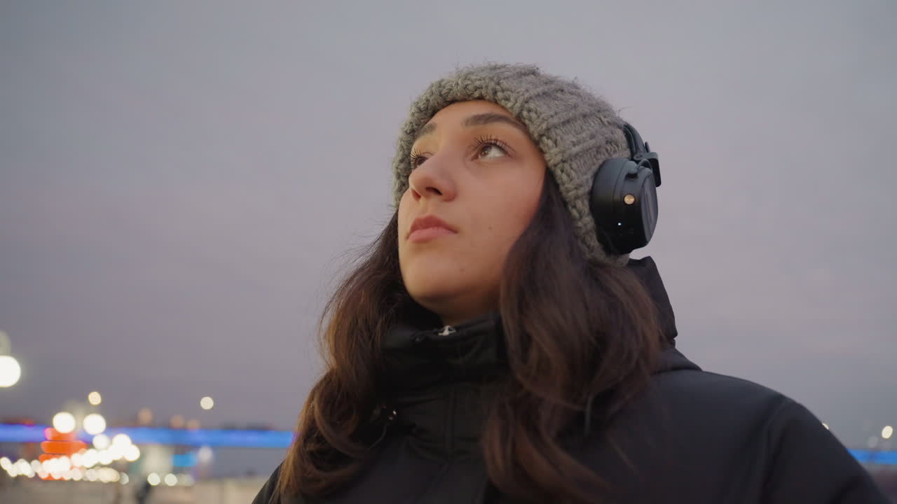 穏やかな夕べの情景。暖かい服装の静かな女性が、夕暮れ時にきらめく街の明かりを見つめている。冬の夜、ヘッドホンをつけた女性が都市の柔らかなイルミネーションを眺める、心安らぐ光景。