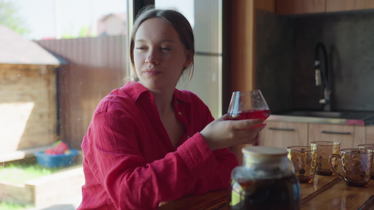 Young woman in red blouse gently sips wine from glass while seated at rustic wooden table near window, surrounded by glass mugs, teapot, and soft indoor lighting