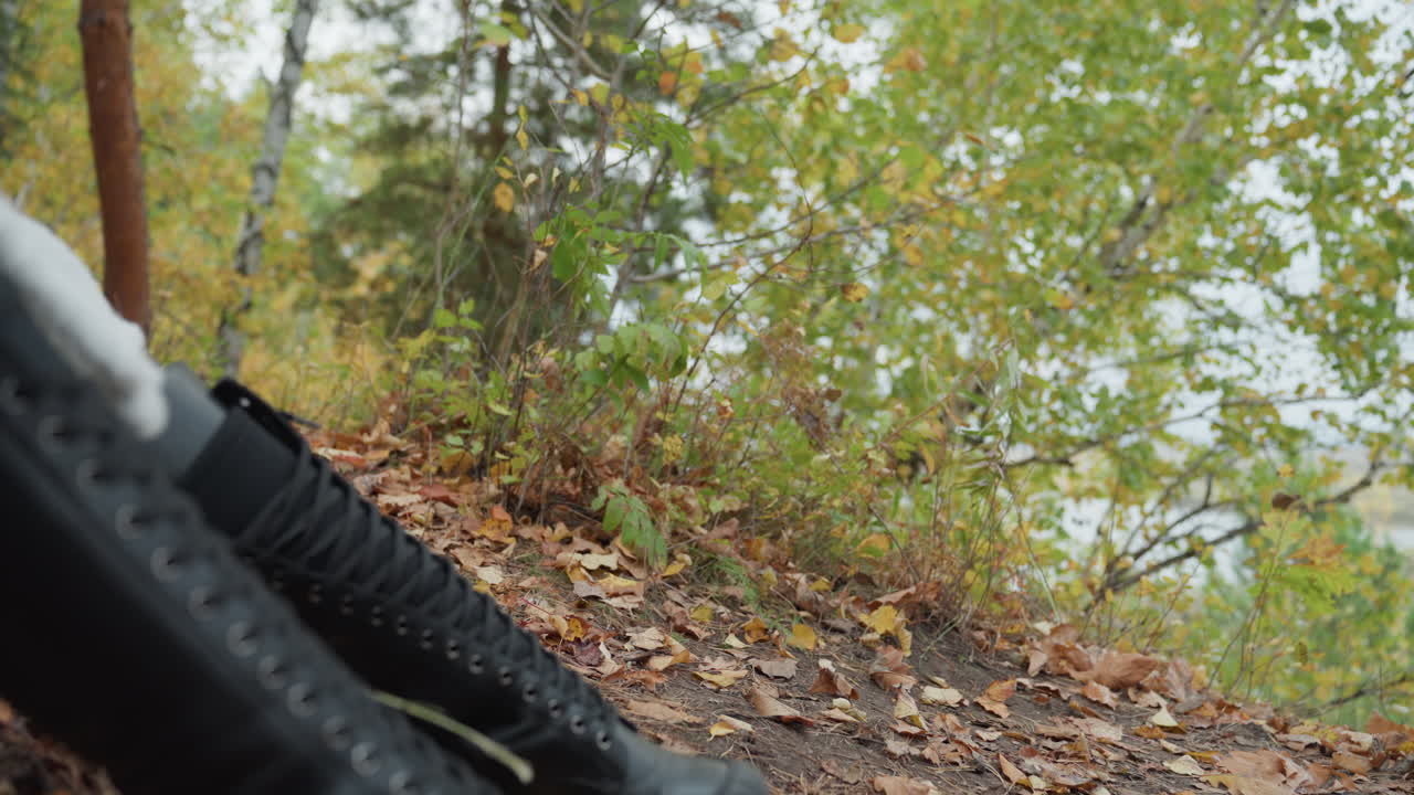 black leather boots on autumn leaves, caucasian woman seated on mossy lakeside bank, tall laceup shafts and chunky platform soles, damp ground and scattered foliage, overcast light, textured
