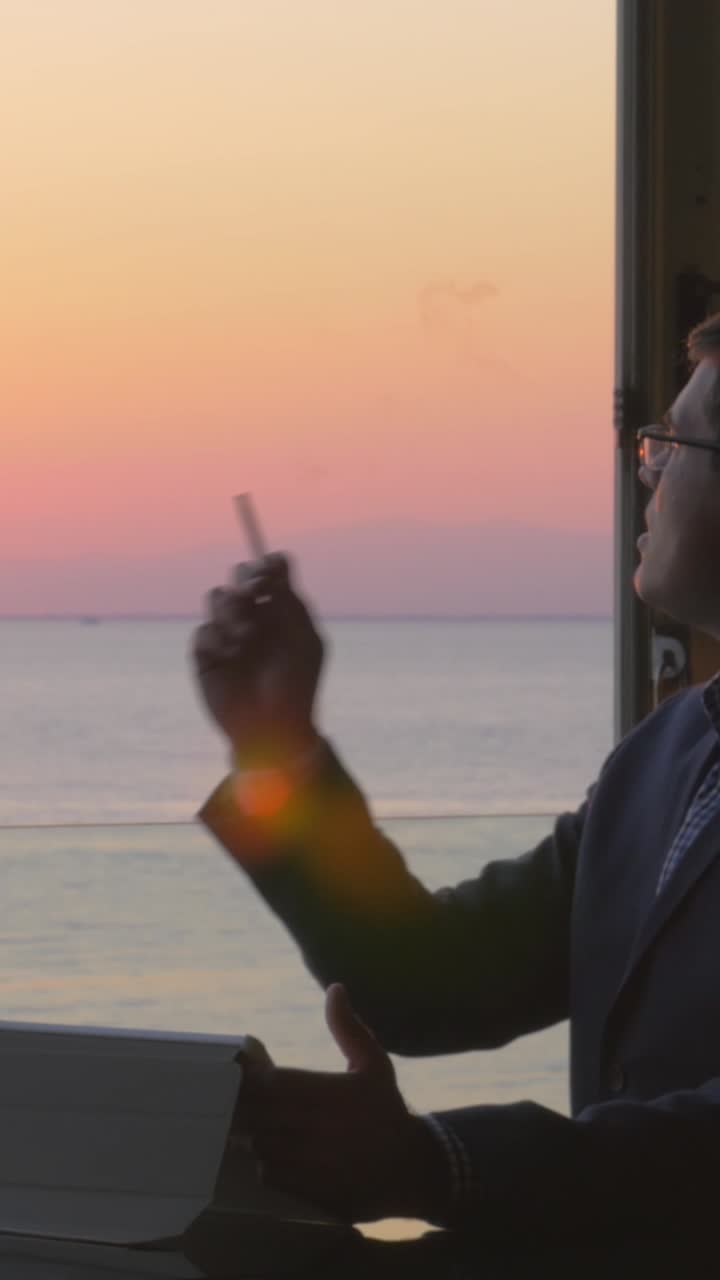 Uomo con occhiali che fuma e usa il tablet in riva all'oceano al tramonto.