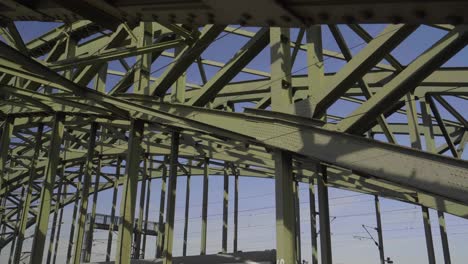 Truck-left-reveal-shot-of-a-commuter-train-from-behind-the-steel-beam-of-a-bridge-looking-up-at-the-architecture-and-sky-with-some-graffiti