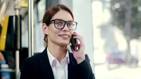 Close-Up-Of-The-Young-Pretty-Businesswoman-In-Glasses-Talking-On-The-Mobile-Phone-With-A-Smile-While-Sitting-In-The-Tram-And-Going-Home