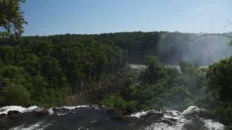 iguazu falls waterfall in argentina, slo-mo waterfall stream falling off huge cliff into distant jungle rainforest landscape, tall greenery surrounding beautiful clear water dropping off cliff edge