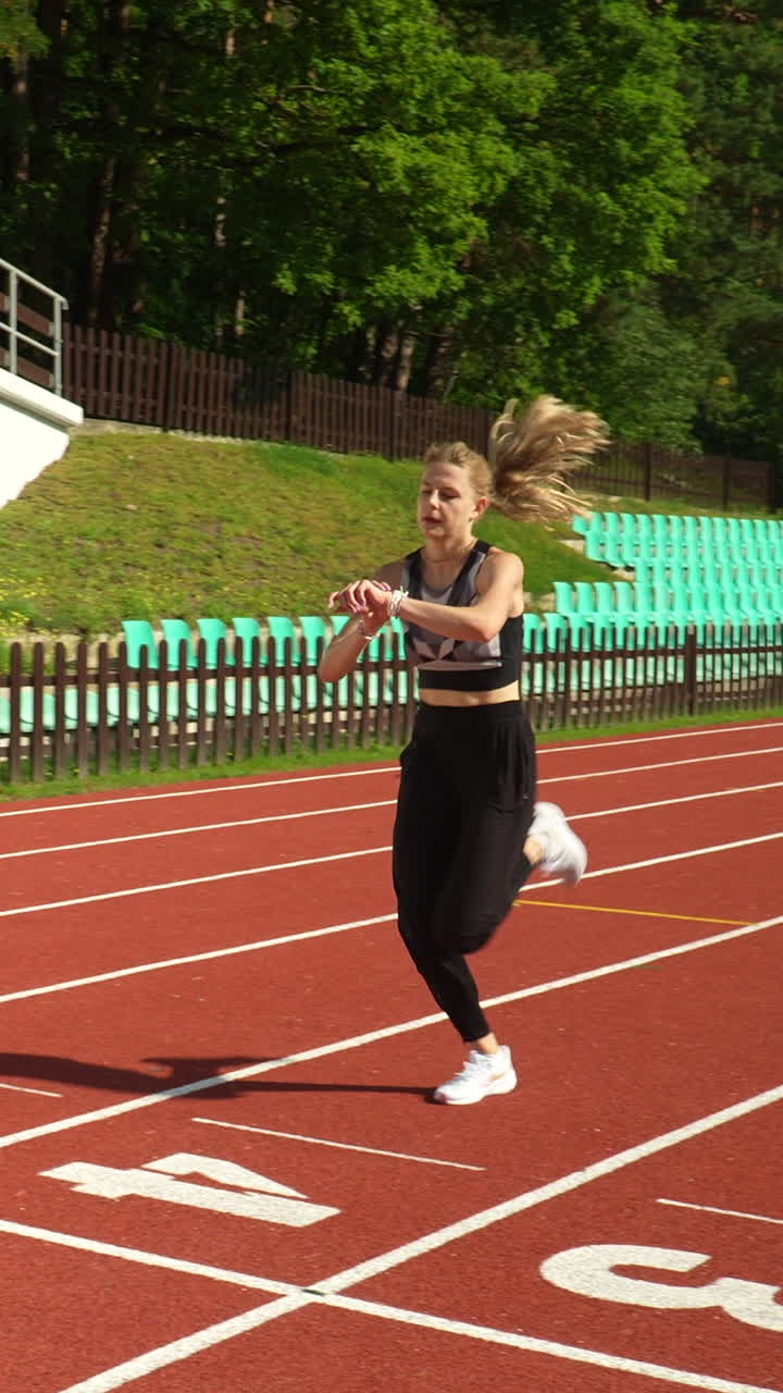 Una ragazza bionda e atletica scatta lungo una pista da corsa rossa in uno stadio all'aperto durante una giornata di sole. Perfetto per app di fitness, storie sui social media e marketing sportivo.