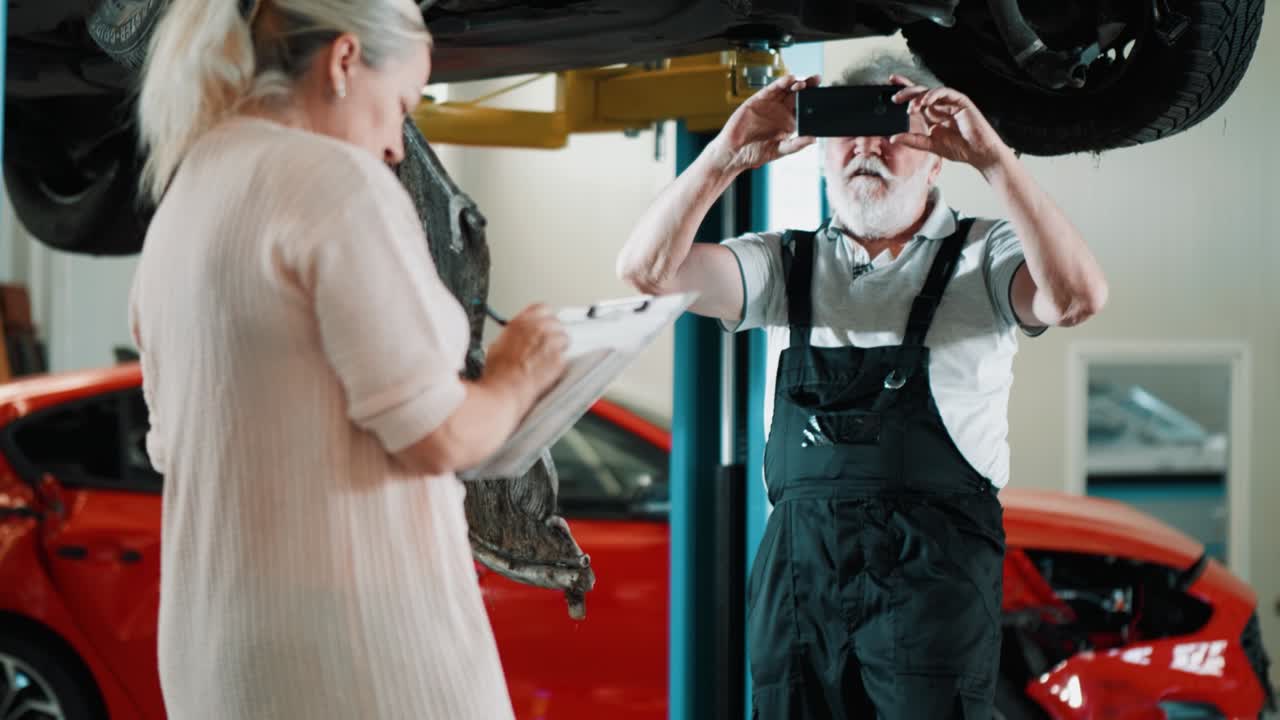Mechanic man making photo a damaged car after a collision, assesses and records the damage in a mechanic's workshop together with a female insurance agent