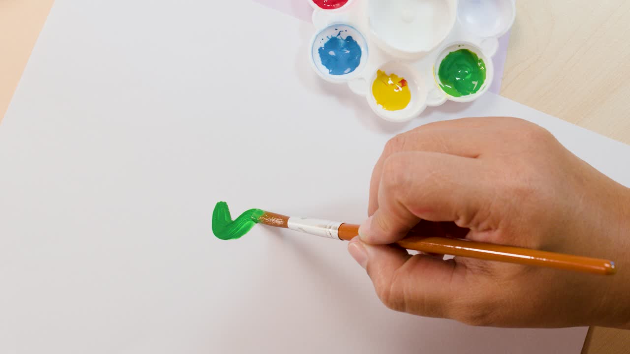 Artist hand paints green wavy line on white paper using brush and acrylic paint palette