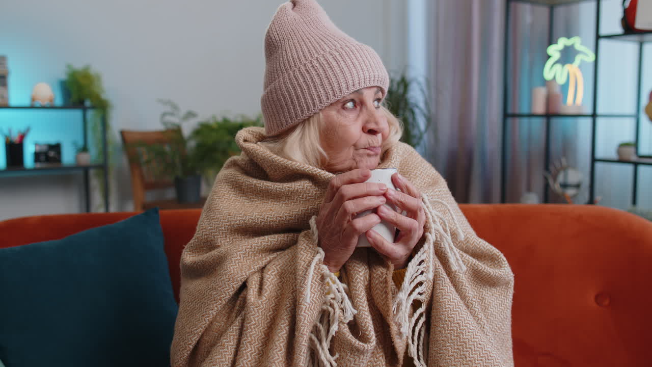 Sick elderly grandmother woman wrapped in plaid sit alone shiver from cold on sofa drinking hot tea