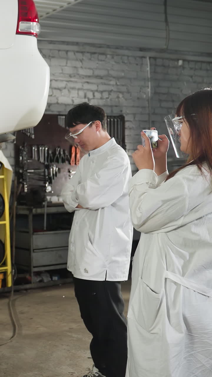 estudiante en bata de laboratorio tomando una foto de un mecánico trabajando en un coche elevado en un taller de reparación de automóviles mientras otro estudiante observa con los brazos cruzados